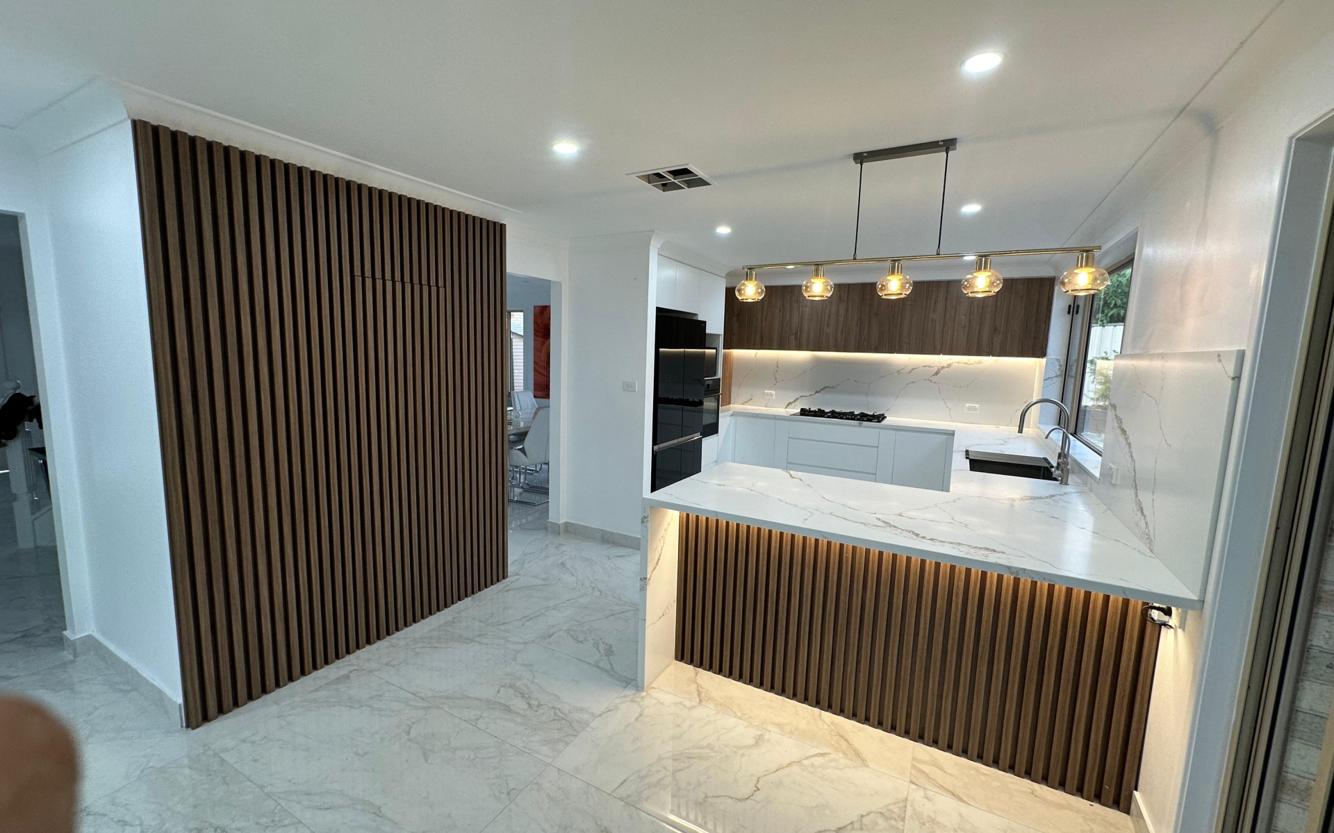 Modern custom kitchen with white marble-look countertops, wood slat feature panels, warm LED lighting, white cabinetry, and a large kitchen island