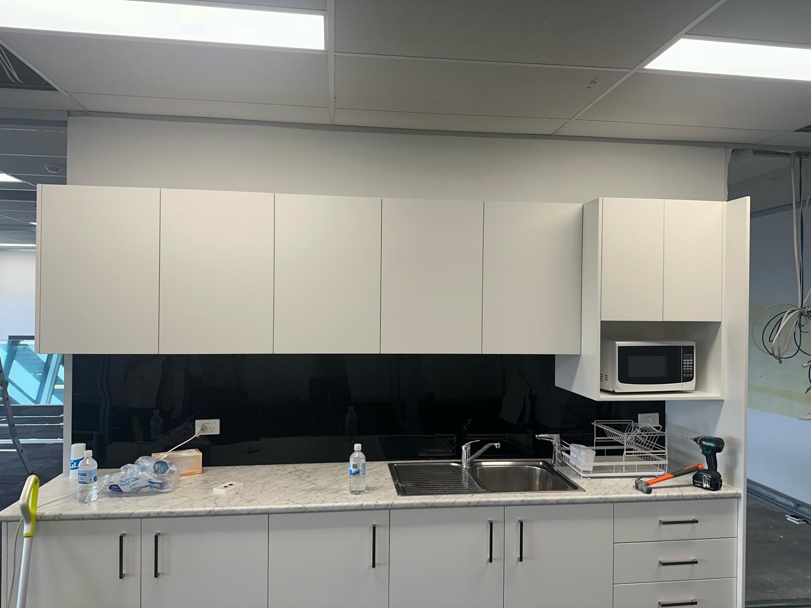 White office kitchenette cabinetry with marble-look benchtop, black splashback and built-in microwave
